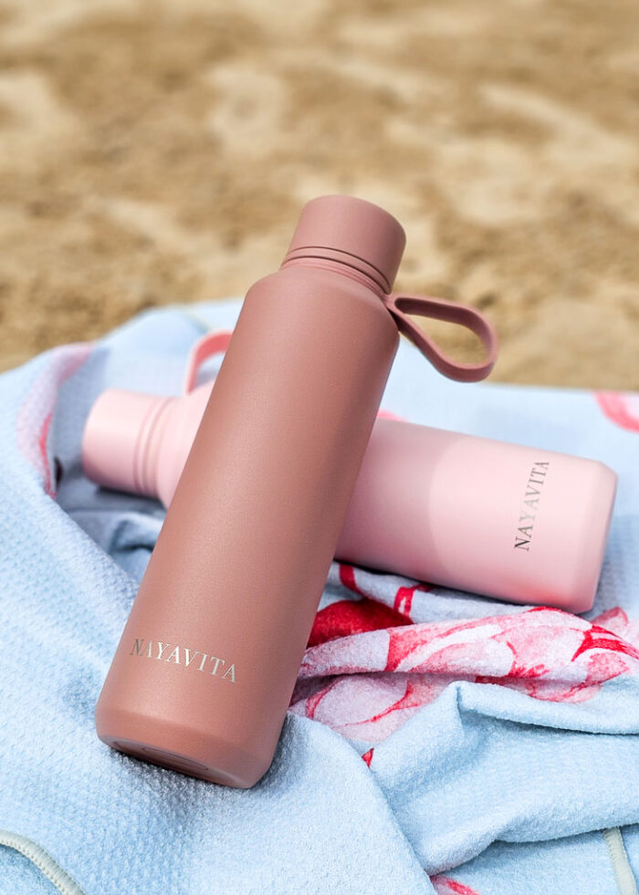 Dvě láhve na vodu NAYAVITA leží na plážové osušce. Láhve mají barvu Terracotta a Pink